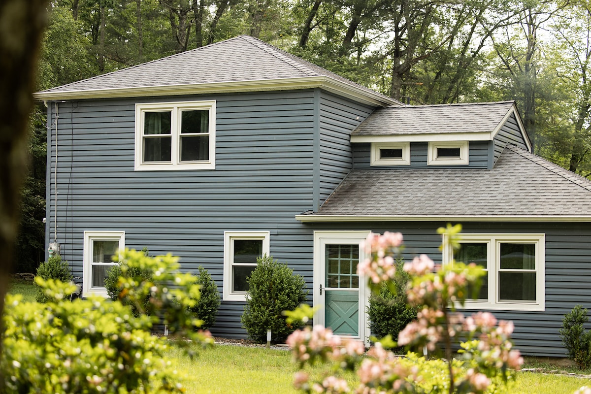 The exterior of the cottage is displayed, featuring a blue facade with multiple windows and a mint green door. Lush greenery and blooming flowers surround the home, enhancing its connection to nature.