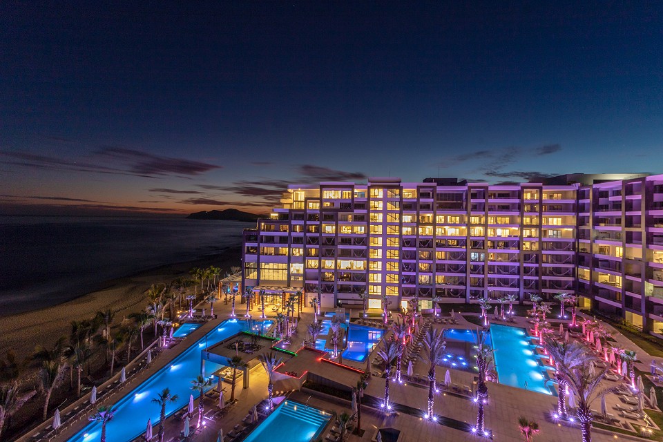 The resort is illuminated at dusk, showcasing multiple outdoor infinity pools with vibrant blue lighting. The modern architectural design of the building features numerous windows reflecting the warm glow from within, while the soft waves of the ocean are visible in the background.