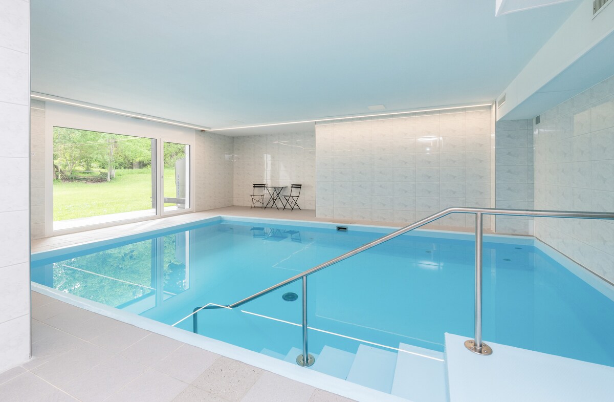 An indoor swimming pool is presented, featuring clear blue water and a smooth tile surrounding. Large glass windows offer views of the greenery outside. A small seating area with two chairs is visible near the pool, enhancing the space's openness.