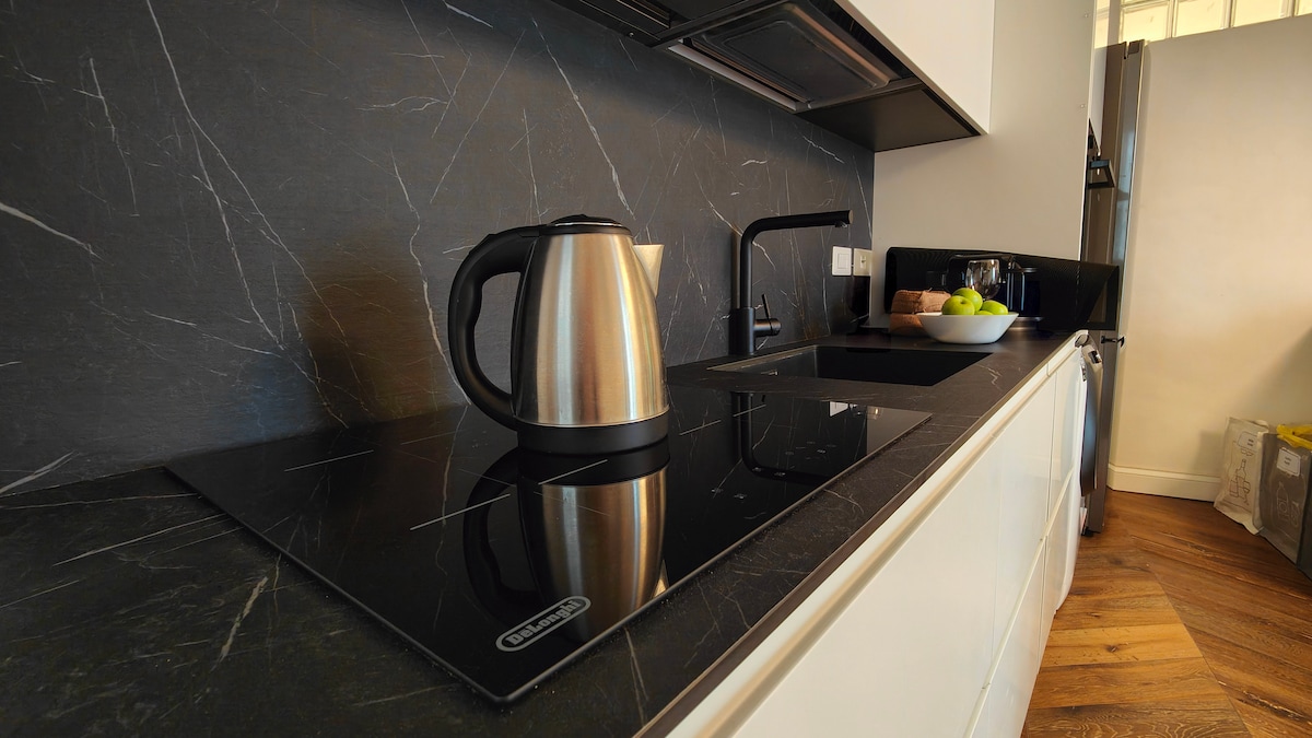 A modern kitchen features sleek black countertops with a minimalist design. A stainless steel kettle sits alongside a glass stove top, complemented by a stylish faucet. Fresh fruit is arranged in a bowl, enhancing the overall functionality and aesthetic of the space.