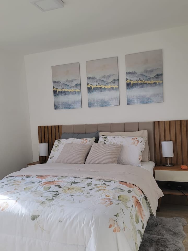 Serene en lichte slaapkamer. Groot bed met zacht beddengoed in bloementinten, voor een diepe rust.
 Houten hoofdeinde met twee lampen, pluizig tapijt aan de voet van het bed. 
Eenvoudig comfort en gegarandeerde rust.