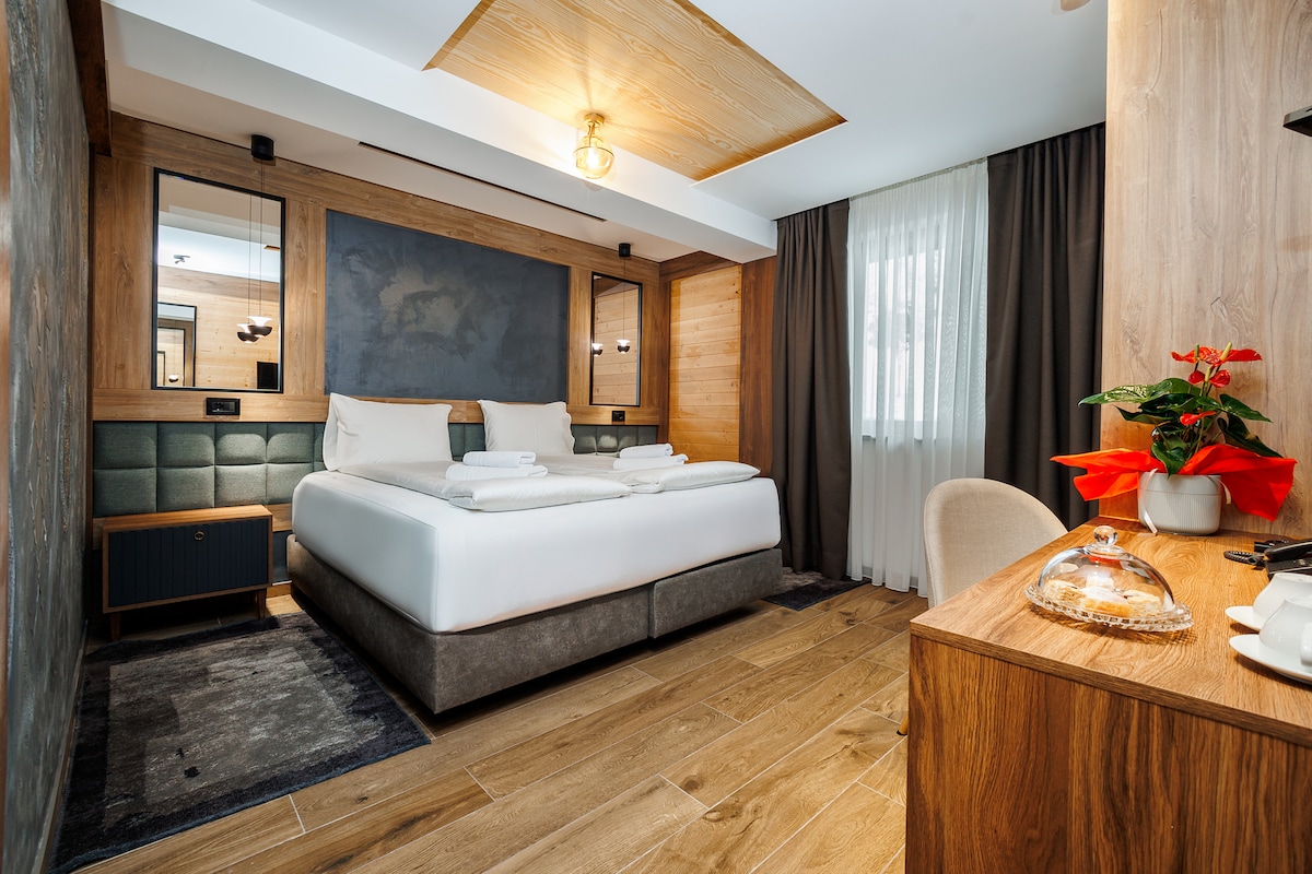 A modern room features a spacious bed with neatly arranged towels and a wooden accent wall. Large mirrors reflect the inviting atmosphere, while a soft area rug complements the wooden floor. A small desk is positioned beside a potted plant, adding a touch of greenery.
