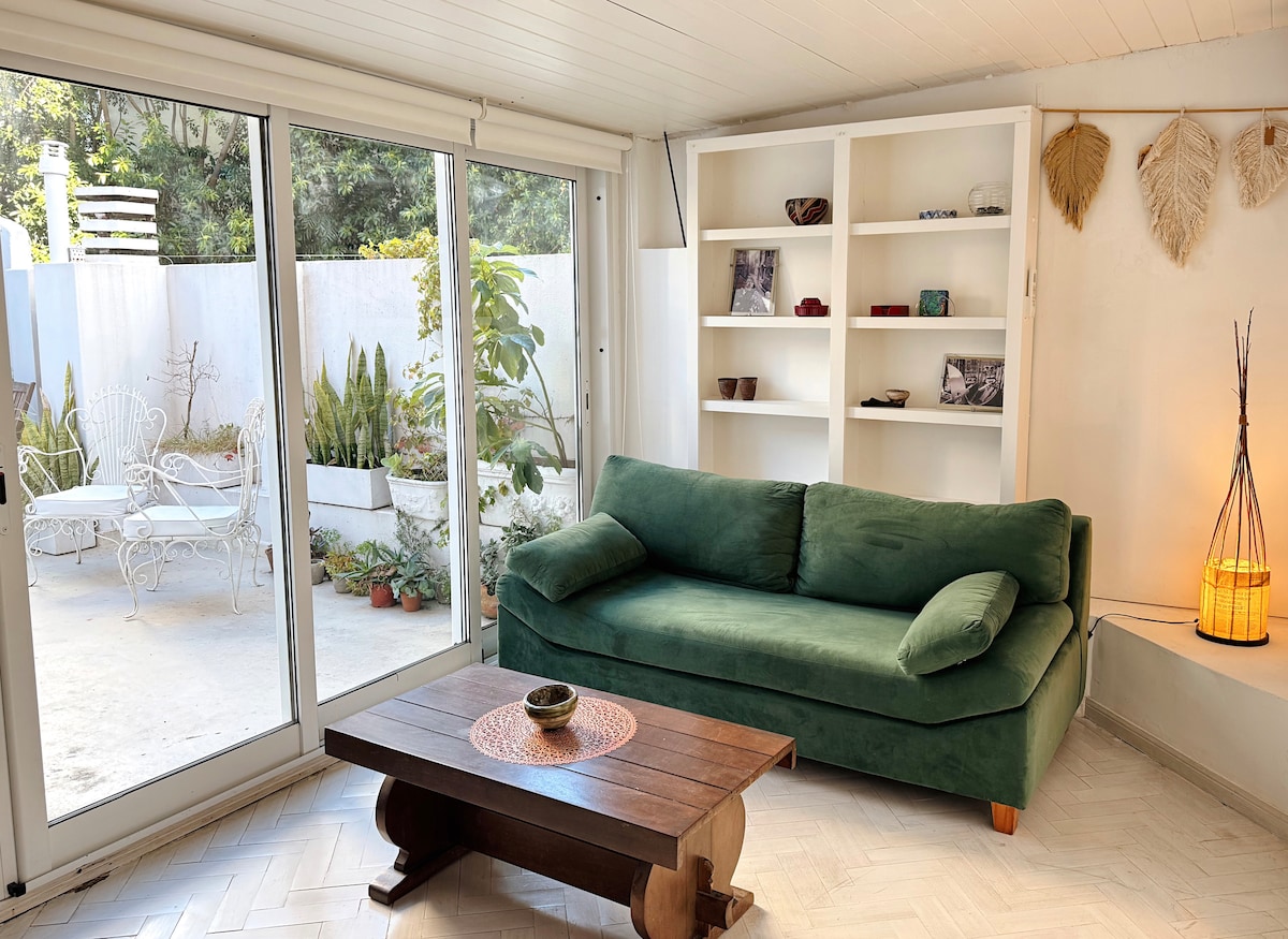 A bright living area features a spacious green sofa and a wooden coffee table, positioned upon a light-colored floor. Large glass doors lead to a tranquil outdoor space with potted plants and a seating area. Natural light fills the room, highlighting the simple decor.