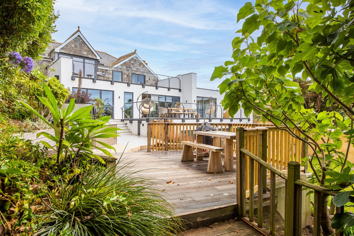 A spacious outdoor terrace provides ample seating with a picnic-style table and benches, surrounded by lush greenery. The modern property is visible in the background, featuring large glass doors that invite natural light into the interior. The setting offers a relaxed ambiance for outdoor gatherings.