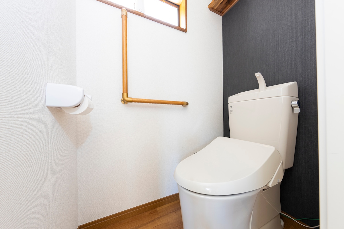A clean bathroom features a modern toilet positioned against a simple wall. A small window allows natural light to brighten the space, while a handrail for assistance is installed nearby for safety.