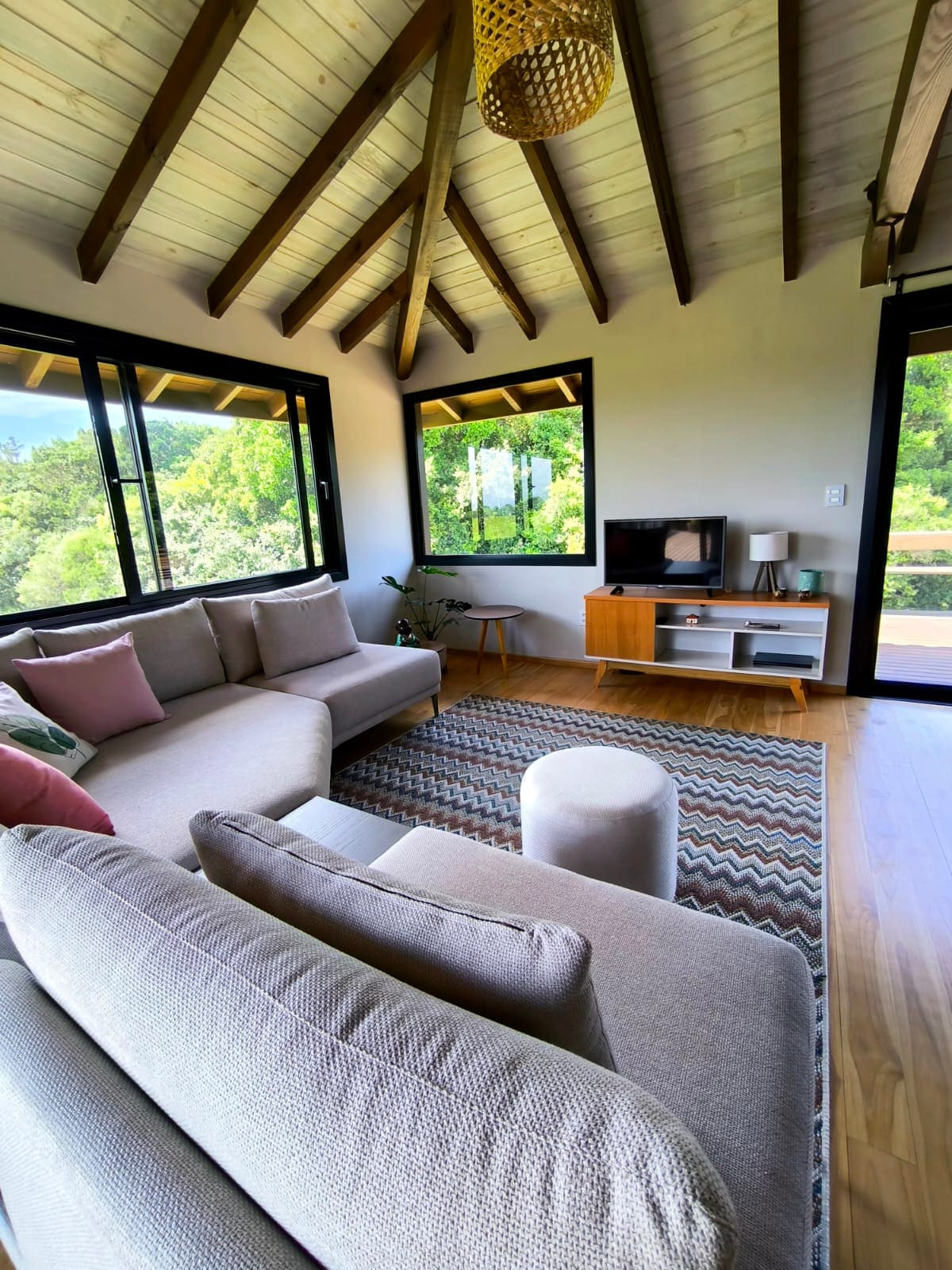 A spacious living area features large windows that frame the natural surroundings. A light-colored sectional sofa is complemented by colorful cushions, while a small side table and a television stand are present. The wood-beamed ceiling enhances the airy feel of the room.