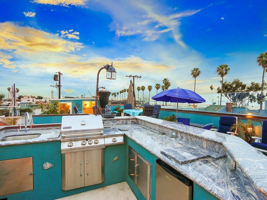 The rooftop deck features a built-in barbecue area with a stone countertop and ample seating, surrounded by palm trees and a clear blue sky. An outdoor umbrella provides shade, creating a relaxing space for enjoying meals and views.