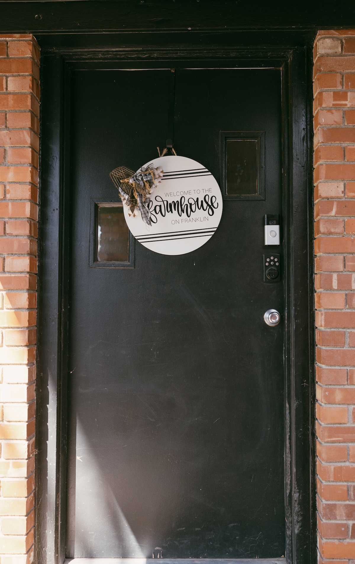 A black front door is presented with a circular welcome sign that reads 'Welcome to the Farmhouse on Franklin.' The door features a small window and a secure lock, while the warm tones of the surrounding brick suggest a charming entryway.
