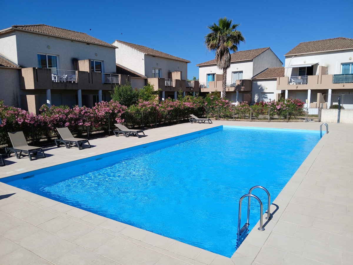 A large swimming pool is surrounded by a tiled deck, featuring several loungers arranged along its edge. Lush greenery and flowering shrubs border the area, while adjacent buildings are visible in the background under a clear blue sky.