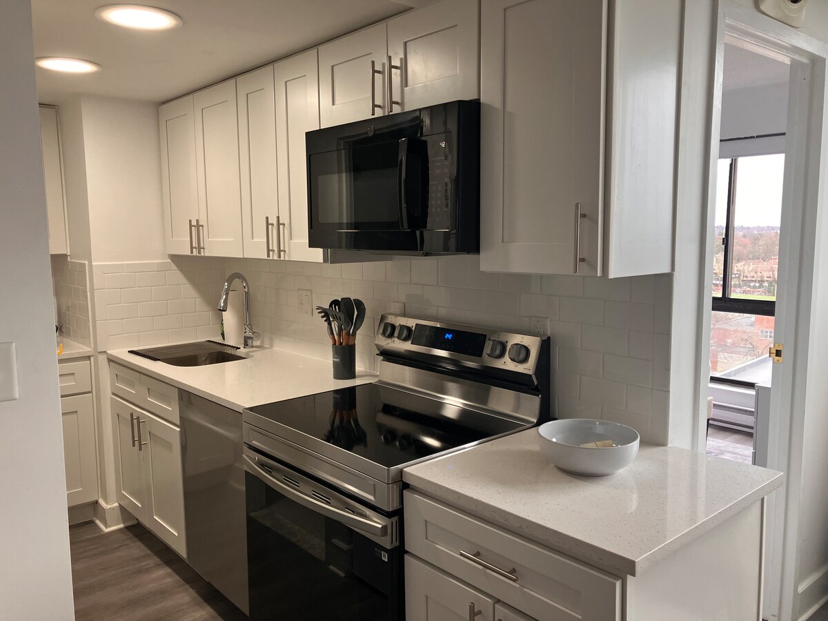 A modern kitchen is presented with sleek white cabinetry and a spacious countertop. Stainless steel appliances, including a microwave and oven, are featured. A bowl sits on the countertop alongside kitchen utensils organized in a holder, enhancing the kitchen's functionality.