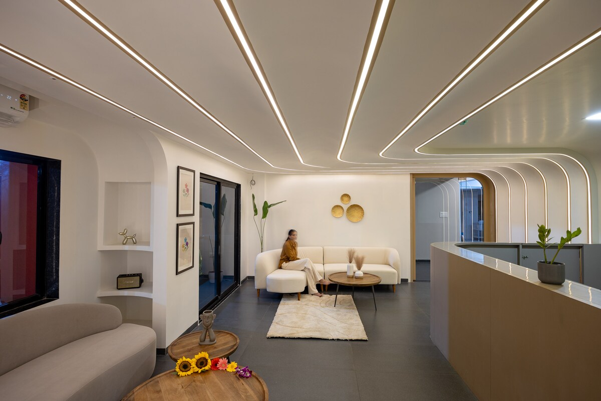 The modern lobby features sleek white walls and a minimalist design. A comfortable seating area includes two light-colored sofas and a wooden coffee table adorned with flowers. Curved ceiling lights and lush plants add a fresh touch to the inviting atmosphere.