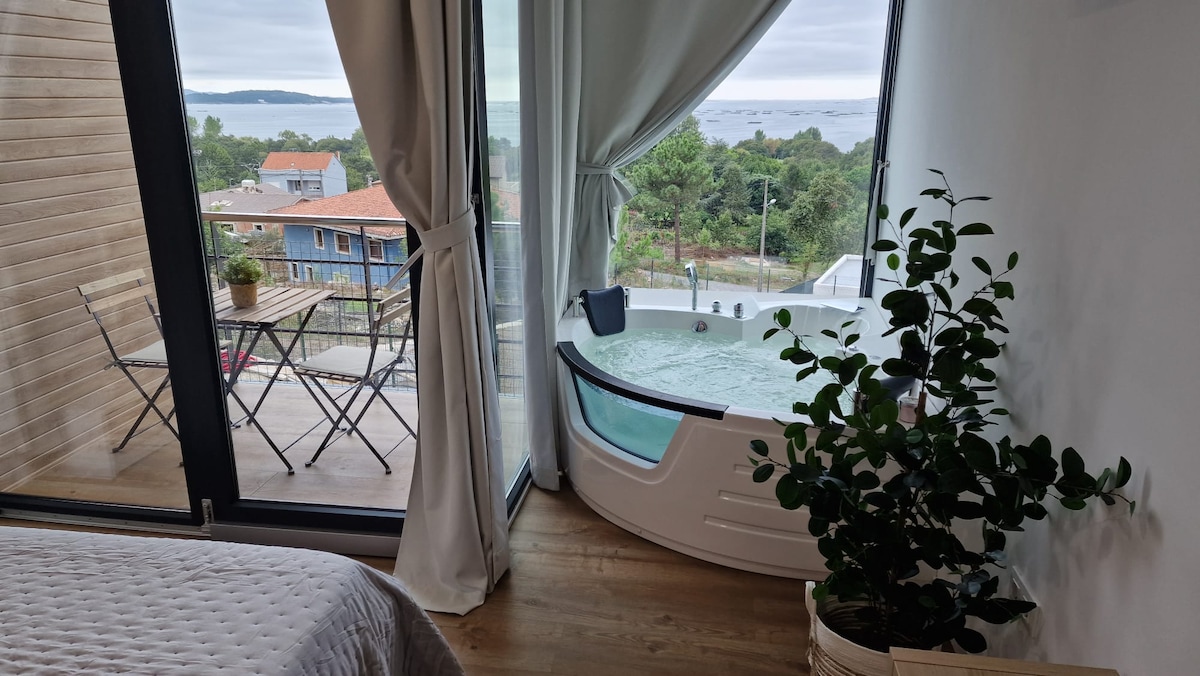 A modern open-concept studio features a private jacuzzi positioned next to a large window, offering views of the nearby bay. A small balcony with seating overlooks the outdoor scenery, complemented by greenery in a potted plant by the window.