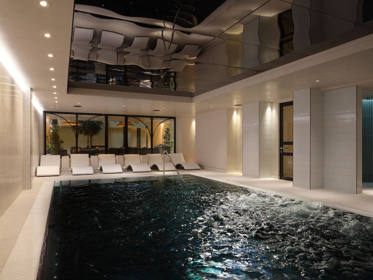 An indoor vitality pool is showcased, featuring a smooth, dark surface with gentle ripples. White lounge chairs are arranged neatly along one side, while large windows provide views of the surrounding greenery. Soft, ambient lighting highlights the sleek tiles and modern design.