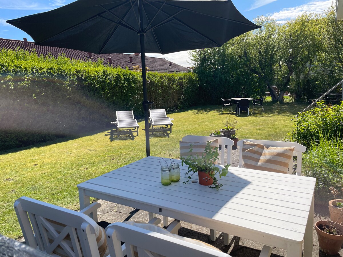 An outdoor dining area features a white table surrounded by chairs, shaded by a large umbrella. In the background, sun loungers and a green lawn are visible, providing a serene space for relaxation. Lush greenery surrounds the area, enhancing the sense of privacy.