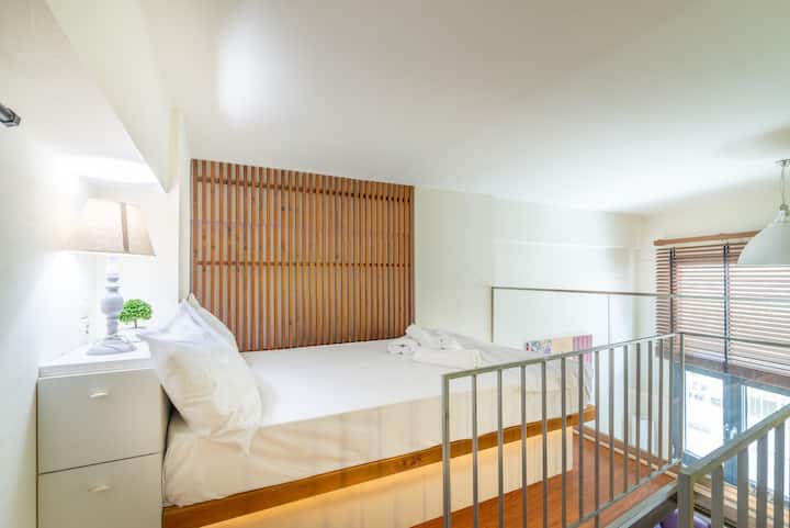 Спокојна спална соба на поткровје со свежа бела постелнина и стилска дрвена табла на глава, која се граничи со модерна метална ограда која додава карактер на дизајнот на поделено ниво.