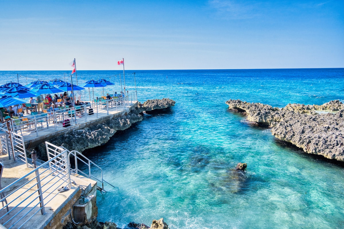 A vibrant scene of the Caribbean Sea is visible, featuring clear blue water and rocky coastline. Umbrella-covered seating areas are positioned on a deck that extends over the water, while flags flutter in the gentle breeze, creating an inviting outdoor atmosphere.