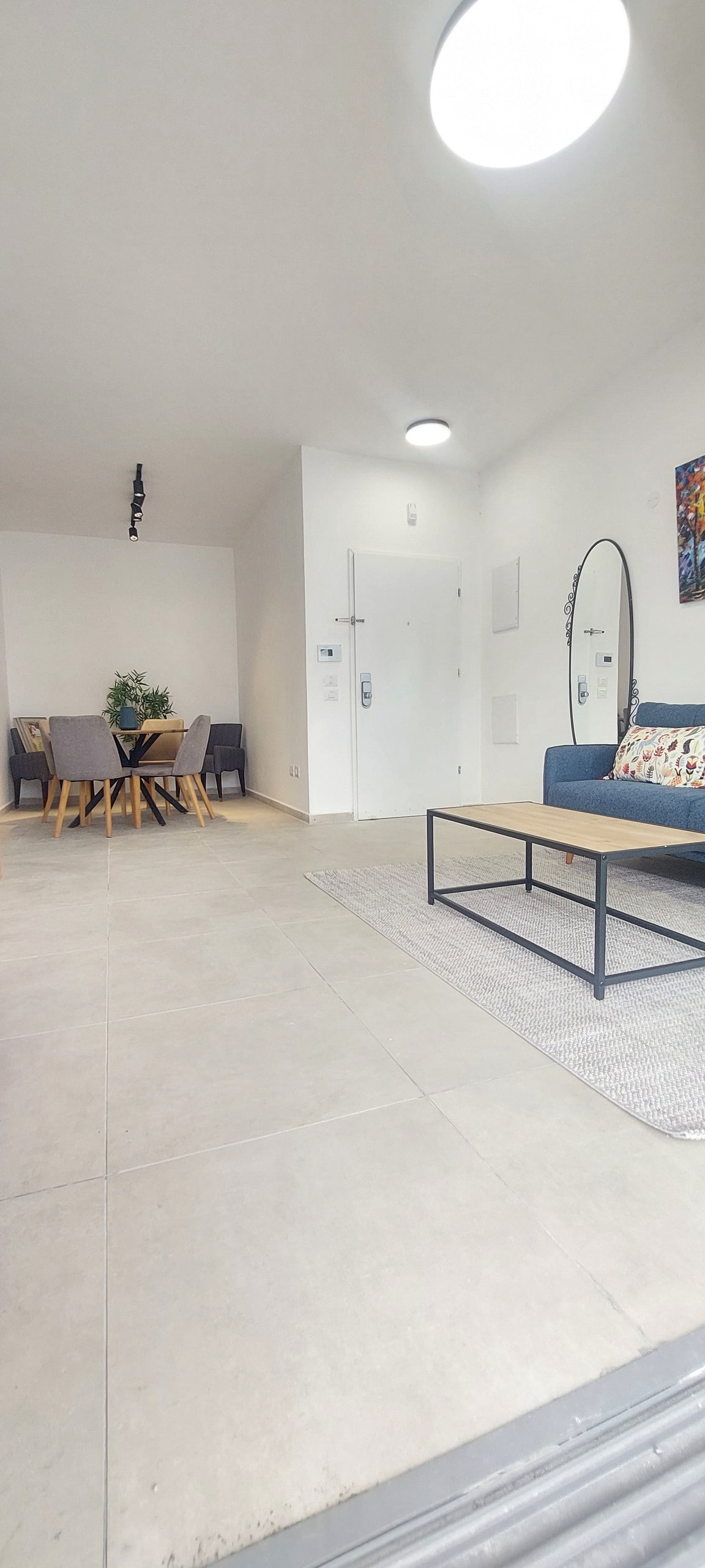 The spacious living area features a comfortable sofa with accent pillows and a wooden coffee table positioned on a soft area rug. A dining table with four chairs is visible in the background, along with a decorative mirror and a piece of wall art. Natural light illuminates the area.