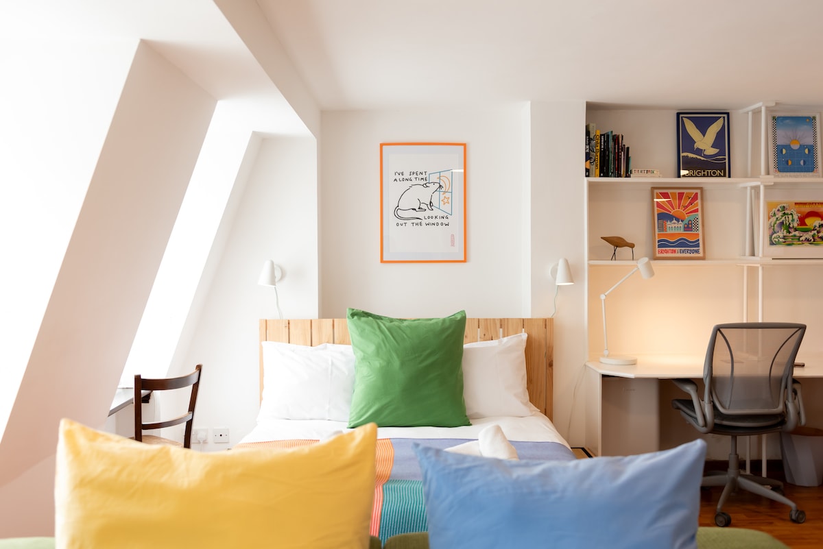 A cozy studio space features a spacious bed adorned with colorful cushions in green and blue. A desk with an ergonomic chair is positioned next to a shelf displaying various art pieces. Natural light fills the room through a window, accentuating its bright, welcoming atmosphere.