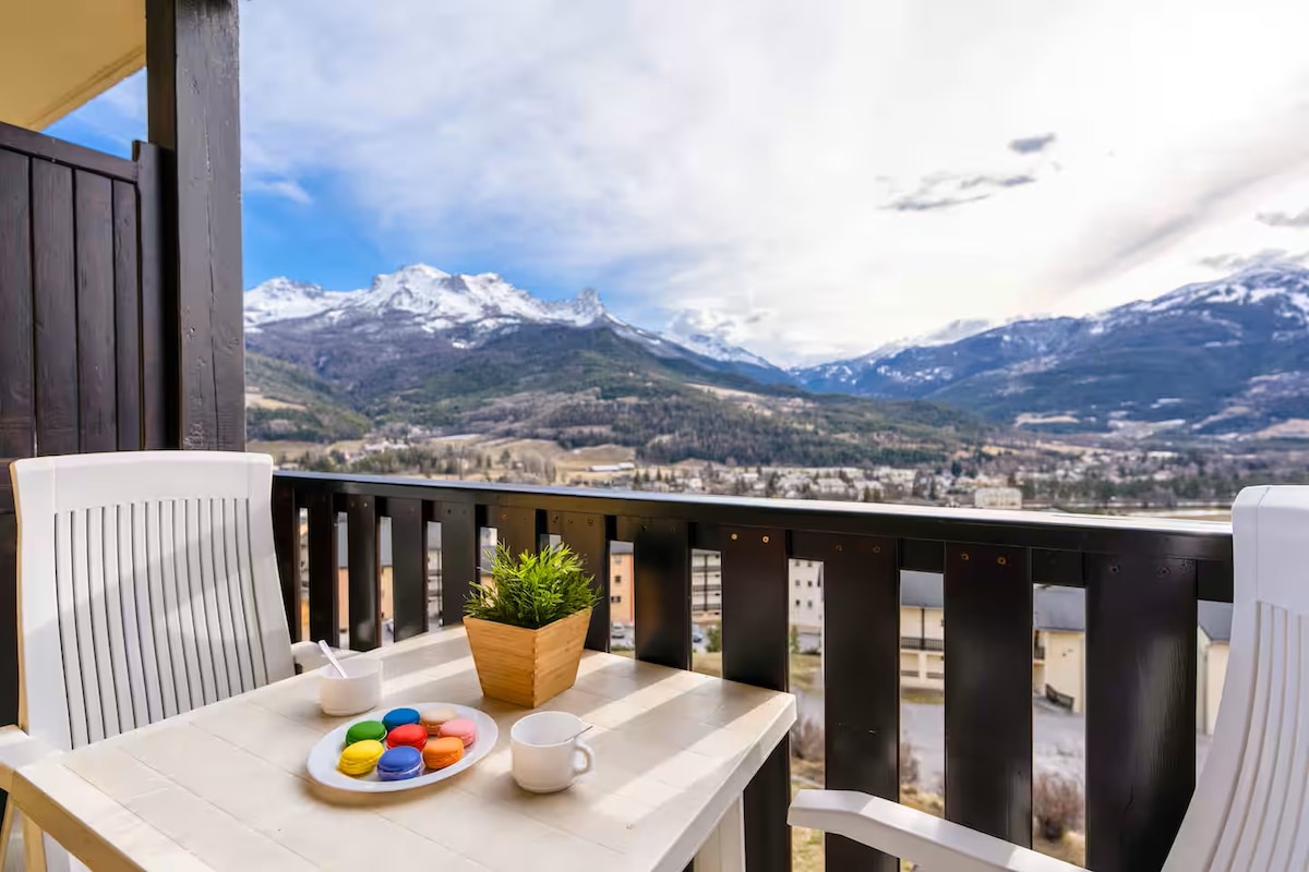A balcony offers a stunning view of the mountains, complemented by a small table and two white chairs. A light-colored pot with greenery sits on the table, alongside a plate of colorful pastries and a white cup, inviting relaxation in a serene setting.