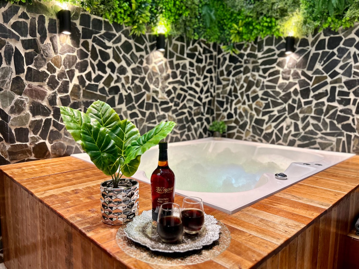 A luxurious jacuzzi is set against a rustic stone wall, with soft lighting enhancing the atmosphere. A wooden table features two elegant glasses filled with dark liquid and a decorative plant in a patterned pot, creating a serene and inviting space for relaxation.