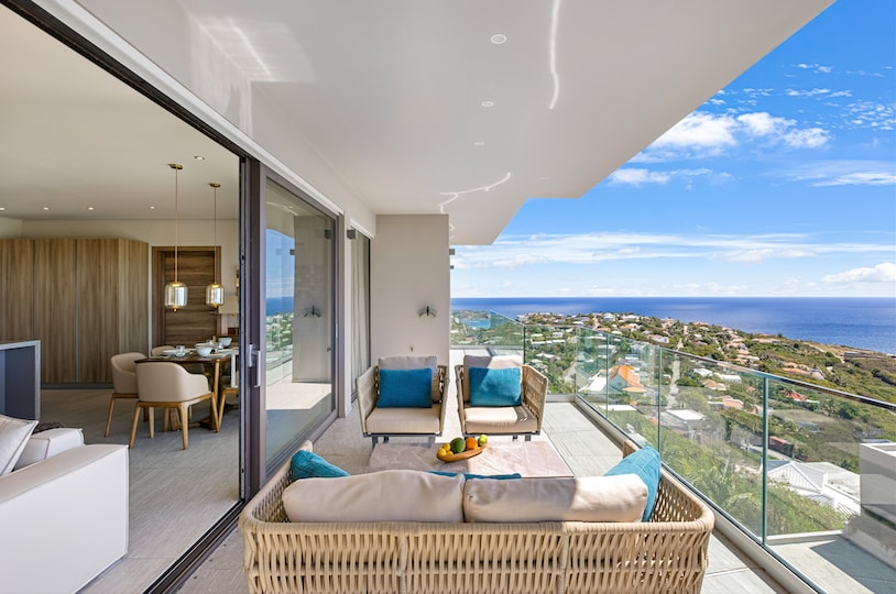 An open terrace features a comfortable seating area with wicker chairs, positioned to showcase views of the Atlantic Ocean and surrounding landscape. Large glass doors connect the terrace to an interior space, allowing for an abundance of natural light.