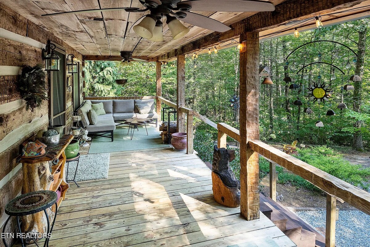 A spacious porch features wooden flooring and rustic logs, surrounded by trees. Comfortable seating is arranged under ceiling fans, while ambient lighting adds to the outdoor charm. Decorative elements, including a colorful mobile and natural sculptures, are visible, enhancing the connection to nature.