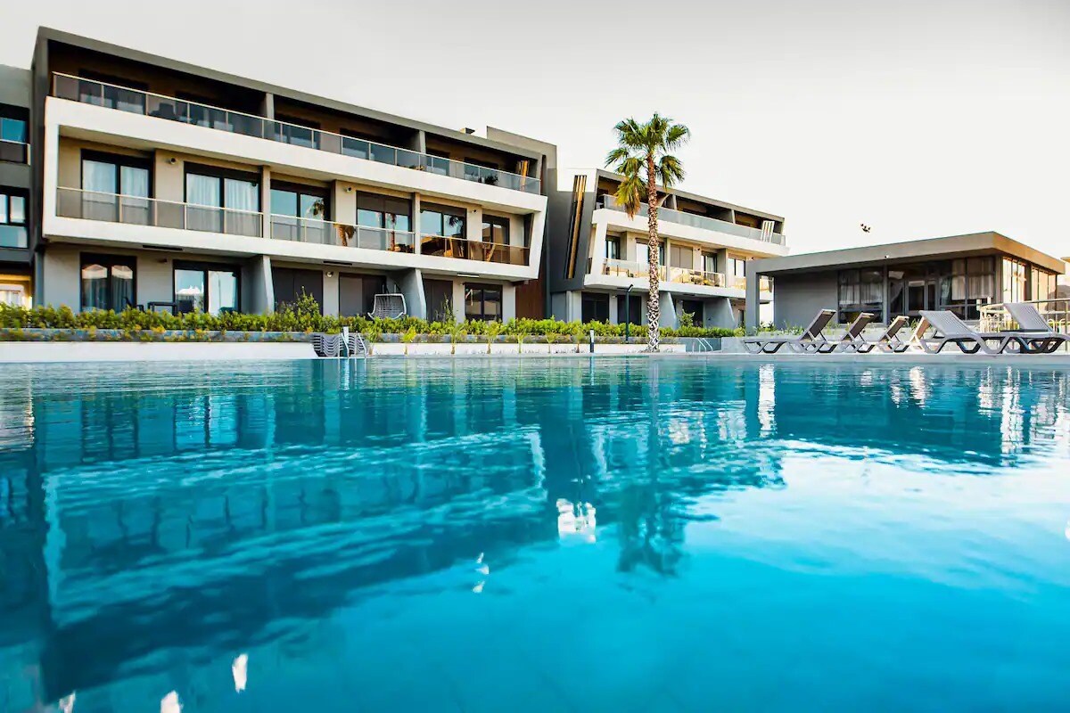 A serene pool area reflects the modern architecture of the surrounding buildings. Comfortable lounge chairs are arranged by the water's edge, while palm trees provide a touch of greenery, contributing to a peaceful atmosphere.