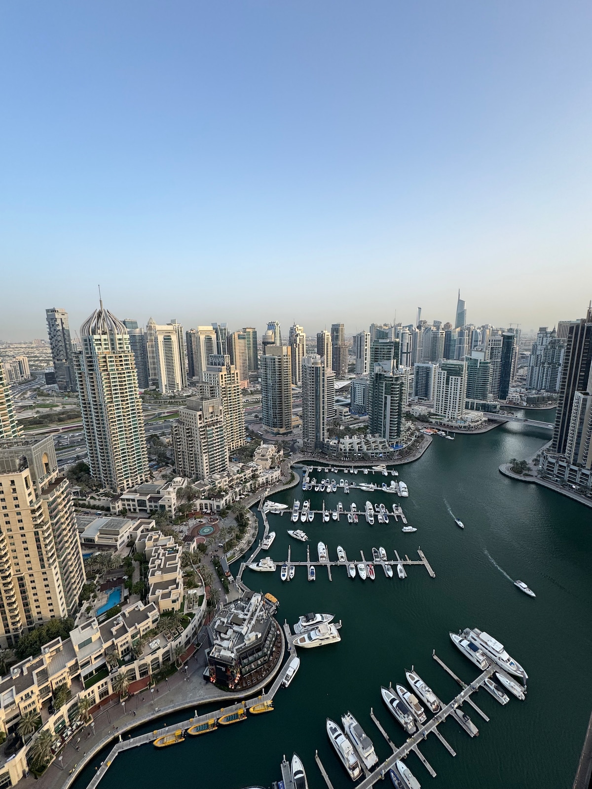 The image captures a panoramic view of Dubai Marina, showcasing a marina filled with numerous yachts and boats. The skyline features a variety of modern skyscrapers, highlighting a blend of architectural styles. Clear blue skies complement the vibrant cityscape, inviting an exploration of the area.