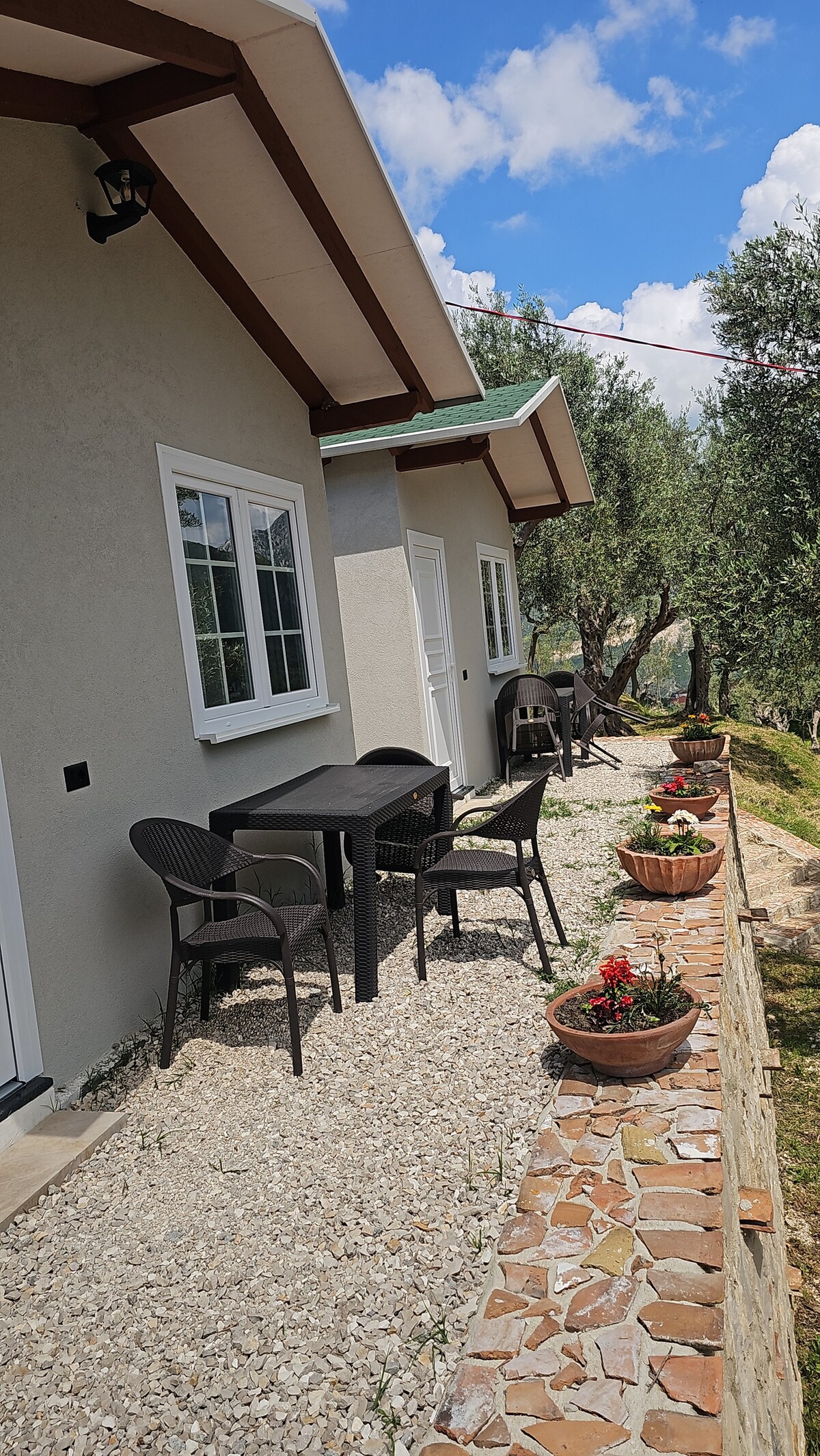 The exterior of a bungalow is depicted, featuring a well-maintained pathway lined with vibrant flower pots. Two sturdy chairs and a table are arranged on a small patio area, surrounded by olive trees and a scenic landscape under a partly cloudy sky.