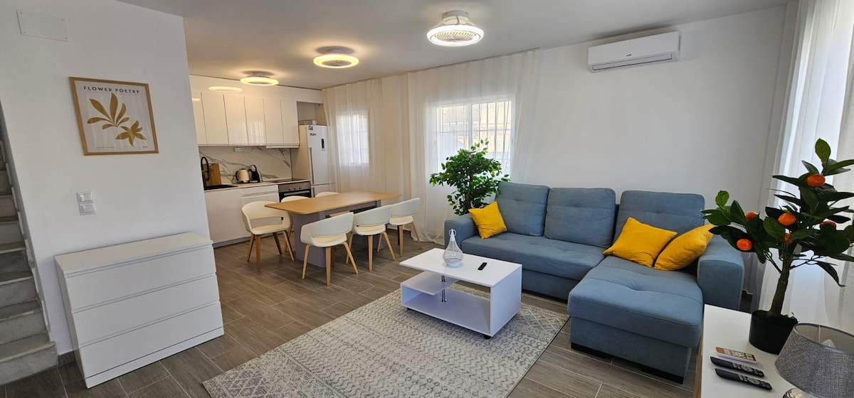 A modern living space features a blue sectional sofa with yellow cushions, positioned next to a white coffee table. A dining area with a light wood table and four white chairs is visible. Natural light enters through sheer curtains, enhancing the clean, minimalist decor.