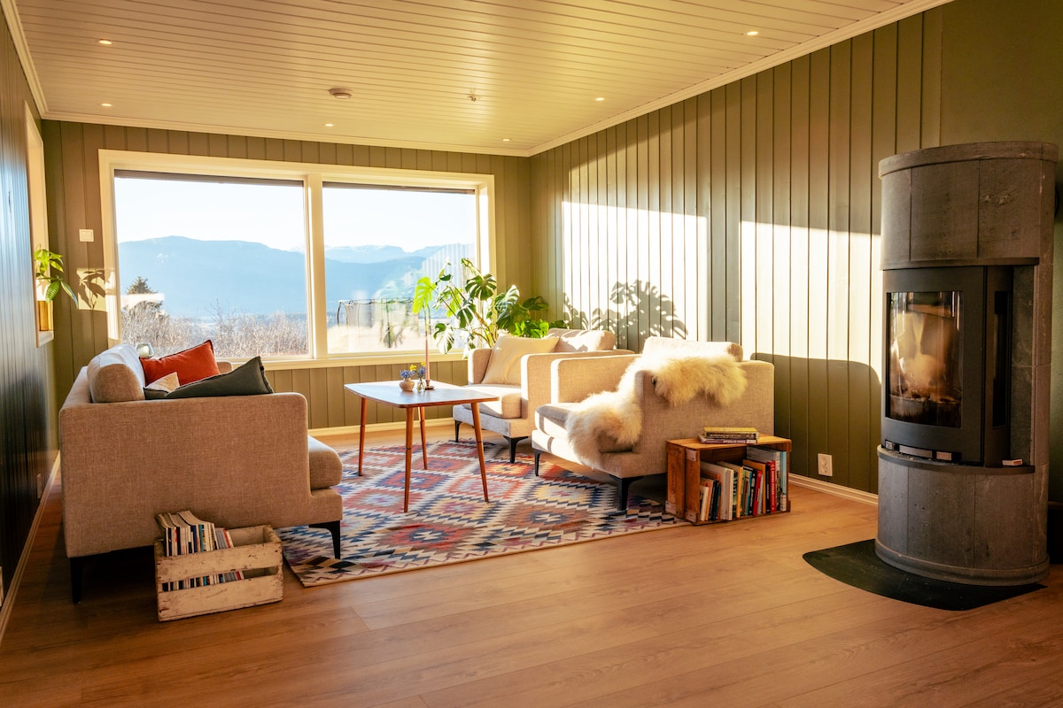 A bright living room is featured with large windows offering scenic mountain views. Comfortable seating is arranged around a central coffee table, with a soft throw placed on one chair. A cozy fireplace stands in the corner, enhancing the space's warmth.