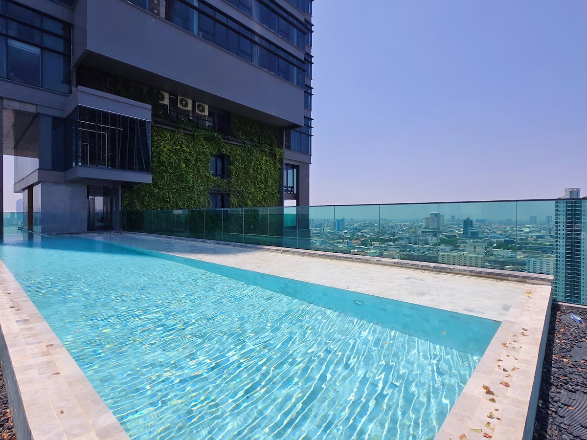 An infinity pool is located on the rooftop, offering panoramic views of the city skyline. The water's surface reflects the sky, and the pool's edge appears to blend with the horizon. Surrounding greenery adds a touch of nature to the urban setting.