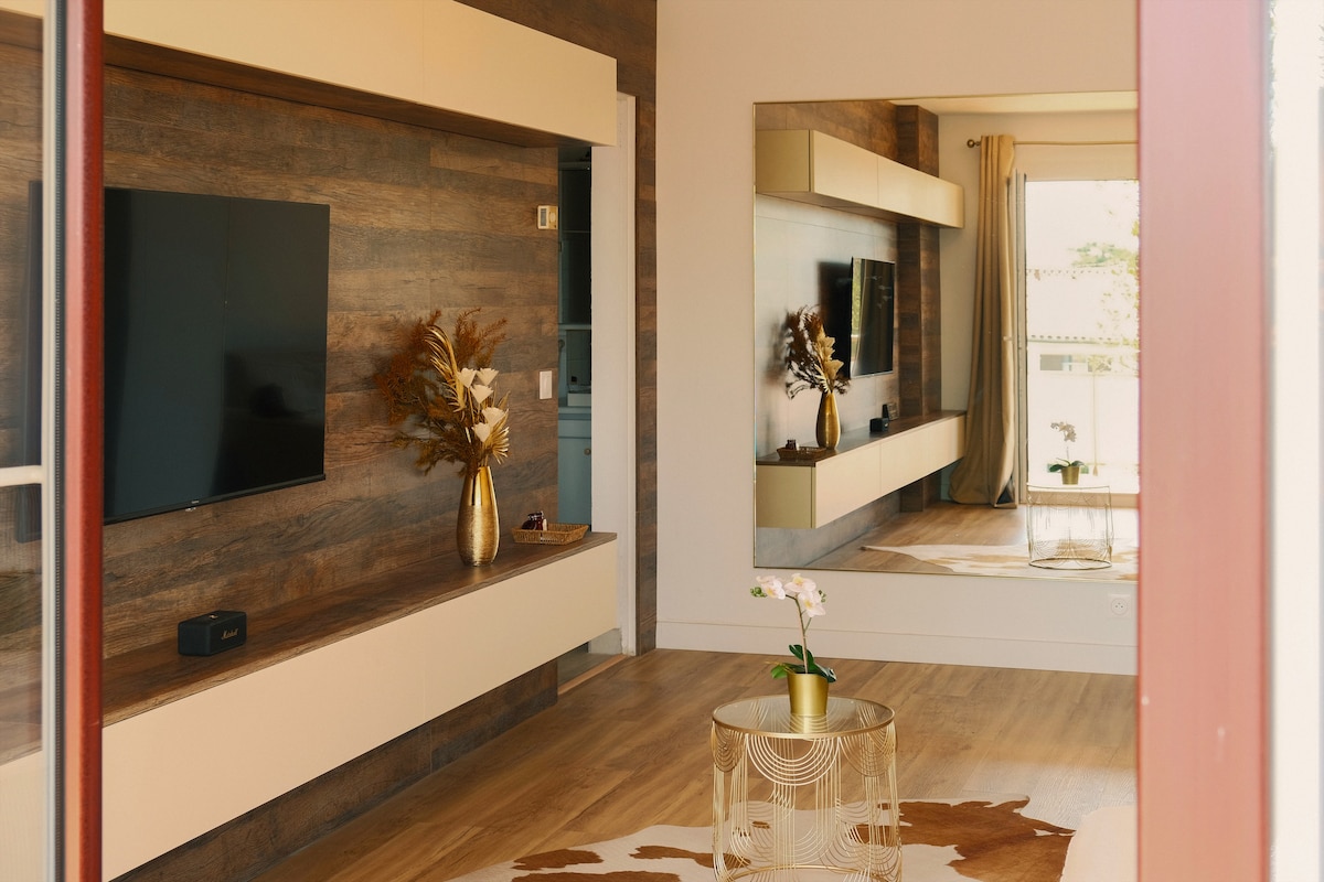 A living area features a large flat-screen TV mounted on a wooden accent wall. A decorative golden vase with flowers is placed on a glass-topped coffee table, complemented by soft lighting and a mix of textures, creating a warm and inviting space.