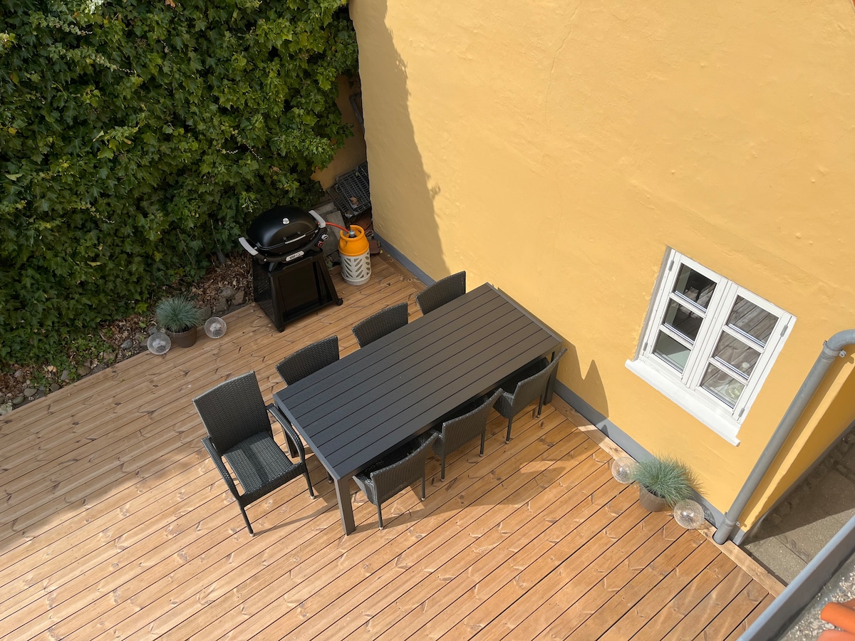 An outdoor terrace features a large rectangular table surrounded by six dark outdoor chairs, set against a backdrop of a vibrant yellow wall. A charcoal grill is positioned nearby, with potted plants adding greenery to the space.