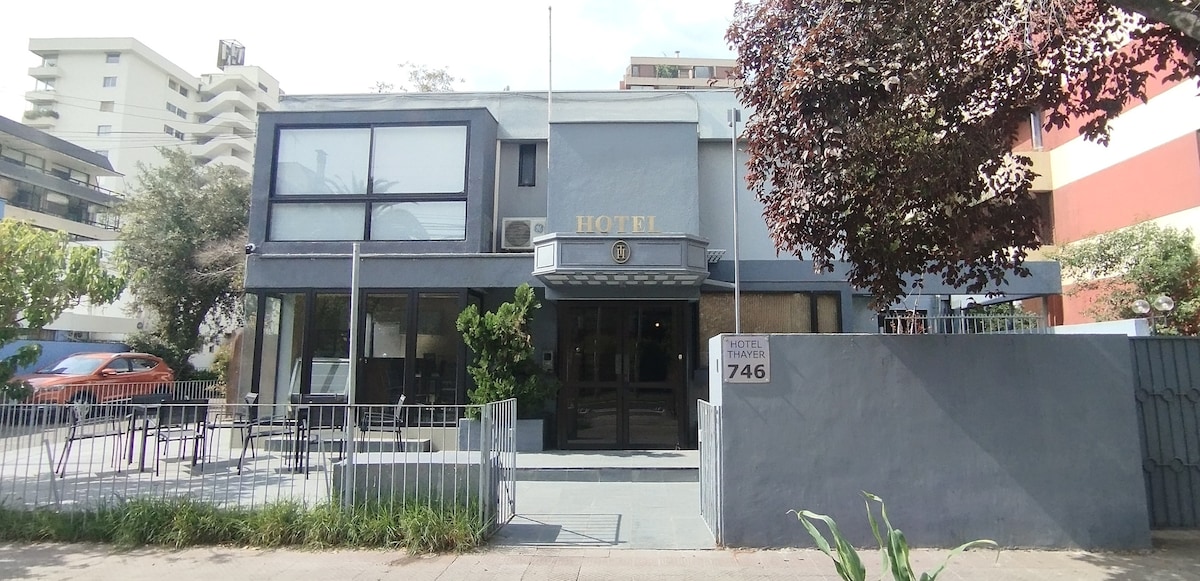 The hotel exterior features a modern façade with large windows and a welcoming entrance. Concrete steps lead to the door, flanked by greenery and potted plants. Outdoor seating is visible on the patio, suggesting a relaxed atmosphere. A gray color scheme complements the contemporary design.