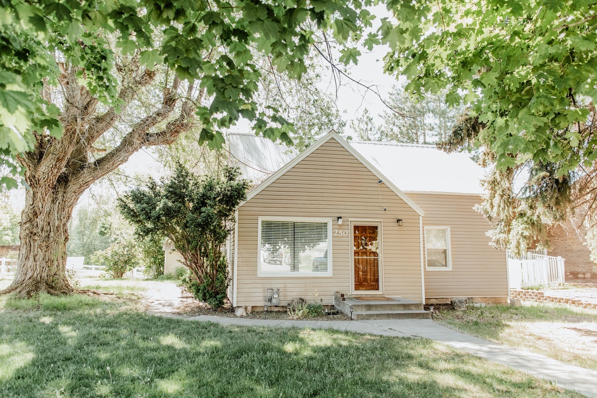 The exterior of a charming two-bedroom home is visible, featuring light brown siding and a welcoming front porch. Lush green trees provide shade, while a manicured lawn borders the walkway leading to the entrance. A large window allows for ample natural light.