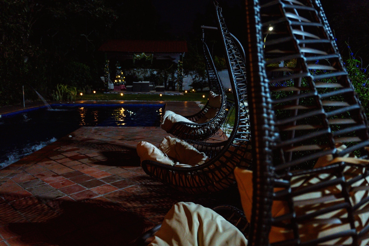 Hanging lounge chairs with cushioned seating are positioned beside a softly lit pool. The tranquil scene is complemented by ambient lighting that reflects off the water, while the surrounding greenery adds to the peaceful atmosphere.