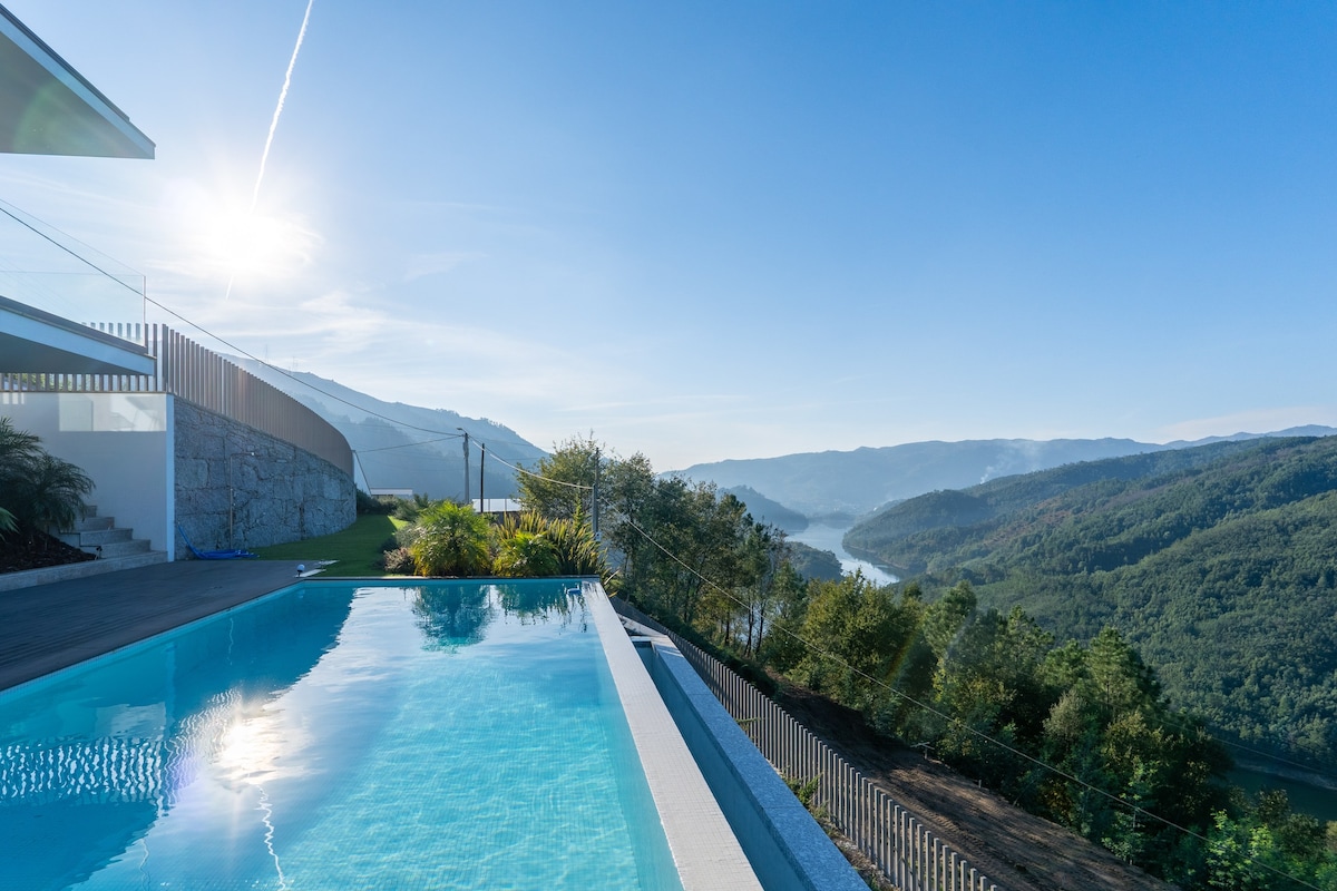 A serene swimming pool reflects the clear sky and surrounding misty hills. A scenic view of a winding river nestled between green valleys is visible in the background, inviting tranquility and connection with nature.