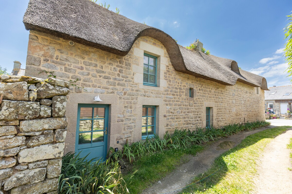 A charming stone cottage with a thatched roof is surrounded by a well-maintained garden. The exterior features light blue windows, providing a contrast against the warm stone walls. A pathway leads up to the entrance, bordered by lush greenery, inviting guests to explore further.
