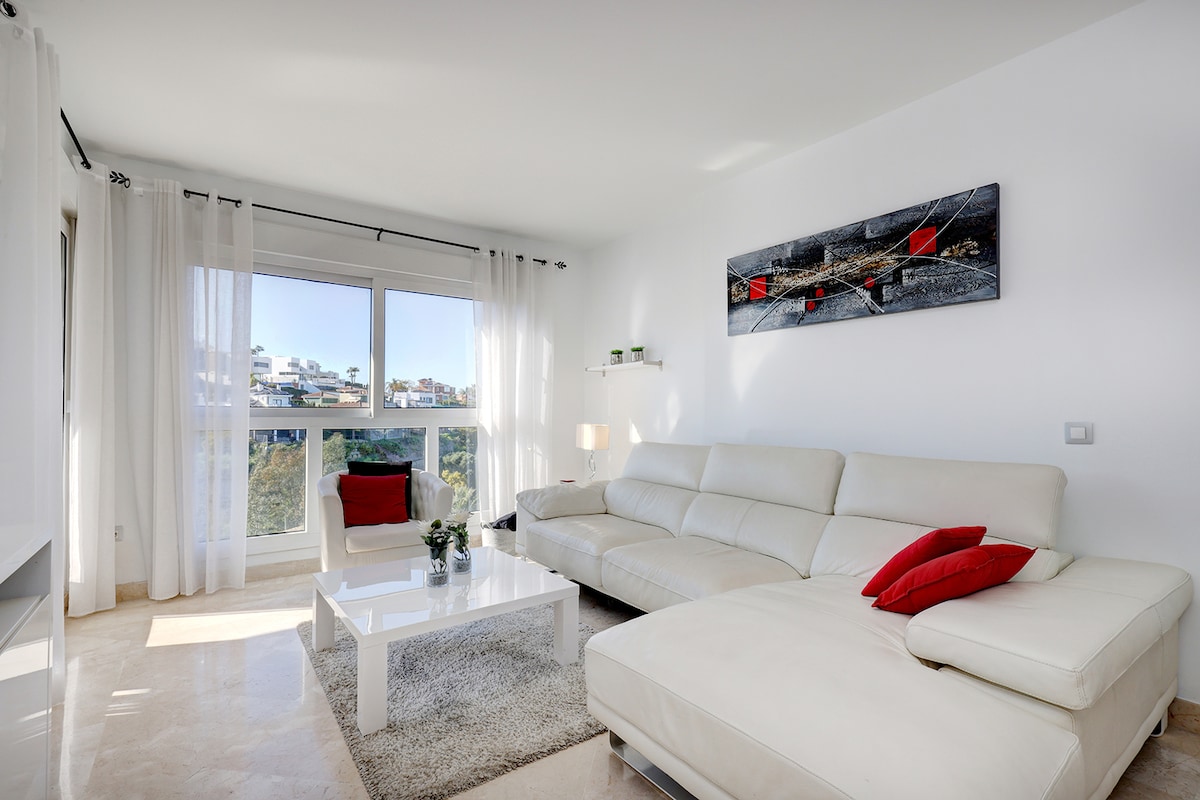 A bright living area features a large white sectional sofa adorned with red cushions. A white coffee table rests on a soft area rug, while large windows provide natural light and views of the surrounding landscape. Minimalist decor is present on the walls and shelves.