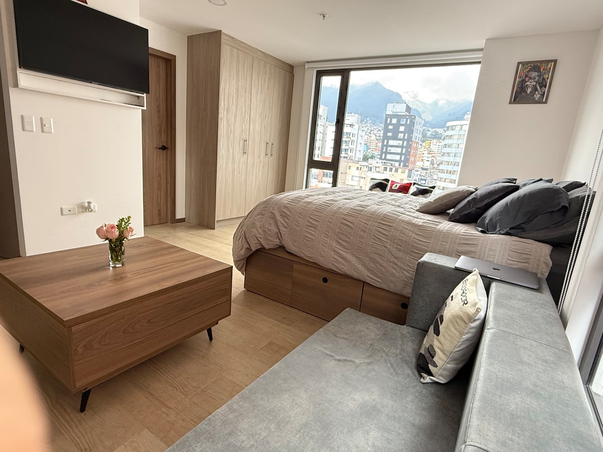 A well-appointed studio is featured, showcasing a spacious bed with light-colored bedding. A modern sofa and a wooden coffee table are positioned nearby. Large windows allow natural light to fill the room, offering views of the surrounding mountains and cityscape.