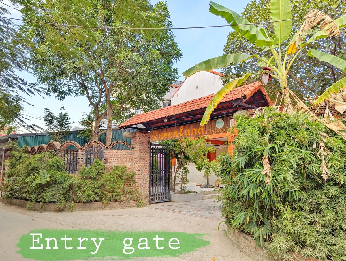 The entry gate is framed by lush greenery, with a rustic brick wall and a decorative metal gate providing access. An overhead tile roof adds charm, while tropical plants and trees enhance the welcoming ambiance of the property.
