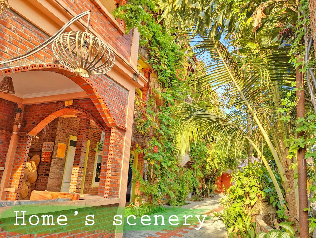 A vibrant outdoor area is framed by lush greenery and flowering vines. A brick structure features arching doorways and warm wooden accents. A peaceful walkway leads to inviting spaces shaded by palms and colorful plants, creating a serene atmosphere for relaxation.