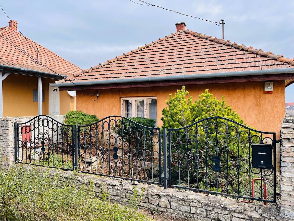 The exterior of the building is presented with a charming orange facade and a traditional red-tiled roof. A decorative black wrought-iron gate frames a neatly landscaped garden with green shrubs and small trees, along with a stone wall that outlines the property.