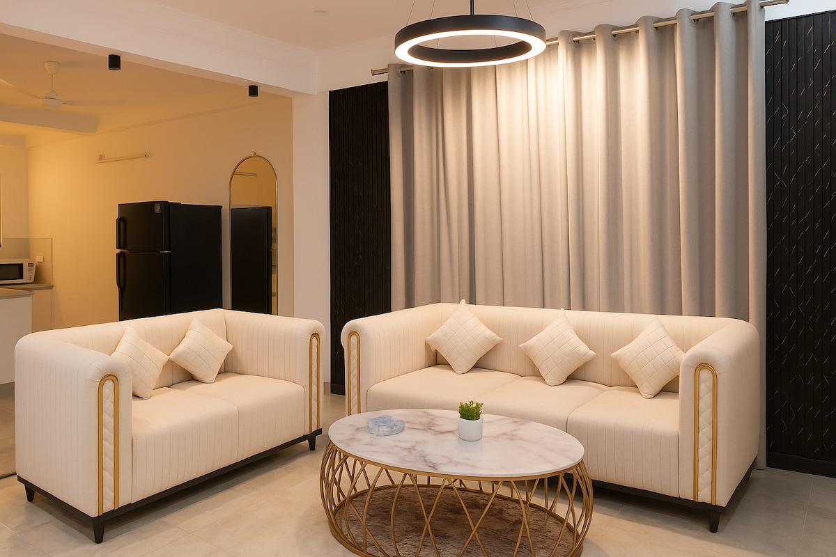 A well-appointed living area is highlighted by two contemporary sofas facing a circular coffee table, set on a textured rug. Soft lighting is provided by a stylish overhead fixture, while the backdrop features elegant drapes and a glimpse of a kitchen area with appliances.