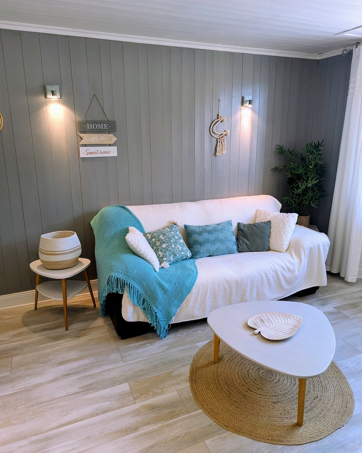 A comfortable living area is presented, featuring a soft sofa draped with a light blanket and adorned with decorative pillows. A small round table sits in front, complemented by a textured area rug. Soft lighting creates a warm ambiance against the gray panelled wall.
