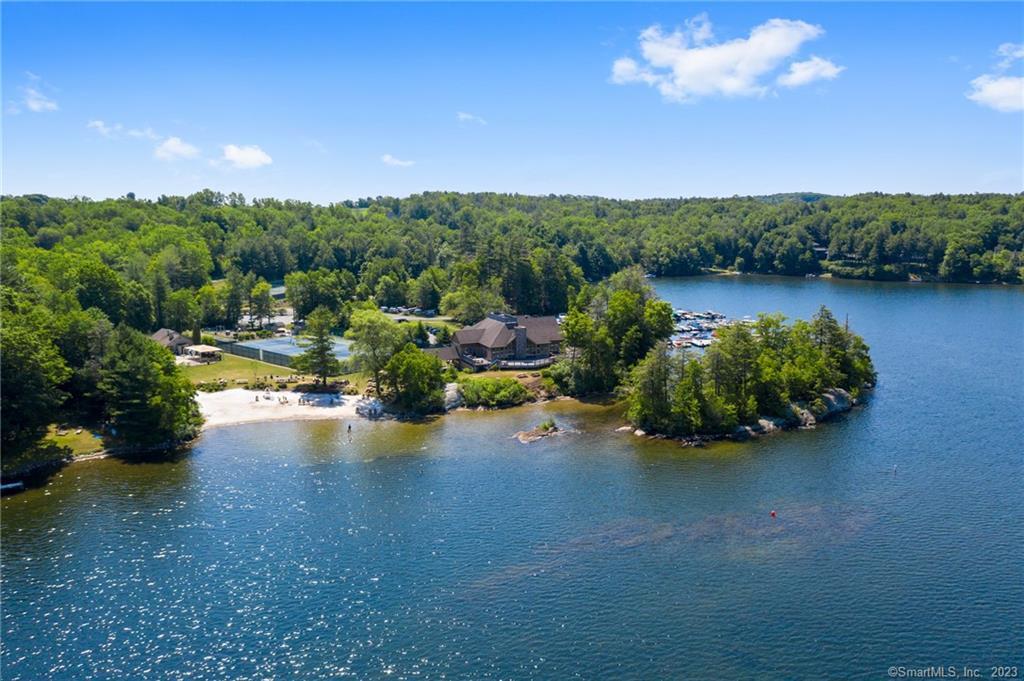 An aerial view of a serene lake community showcases lush greenery surrounding the water. A beach area is visible, along with recreational facilities. Several boats are anchored nearby, and the landscape features a mix of trees and open spaces, creating a tranquil setting.