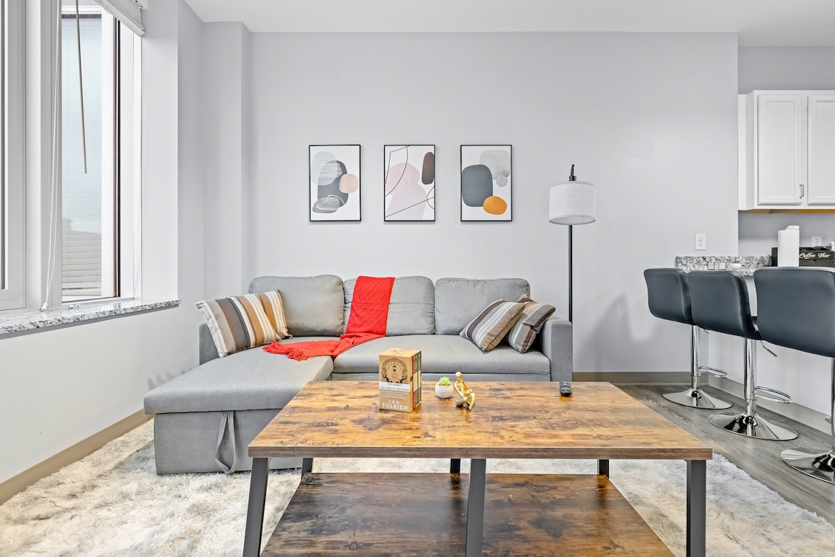 A contemporary living area features a gray sectional sofa adorned with colorful throw pillows and a red blanket. A wooden coffee table sits in front, accompanied by decorative items. Large windows allow natural light to fill the space, enhancing the modern aesthetic.