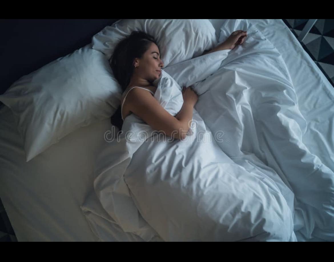 A spacious bed is surrounded by plush, white bedding, featuring a soft duvet and multiple pillows. The tranquil setting is complemented by a dark wall in the background, enhancing the sense of comfort and relaxation.