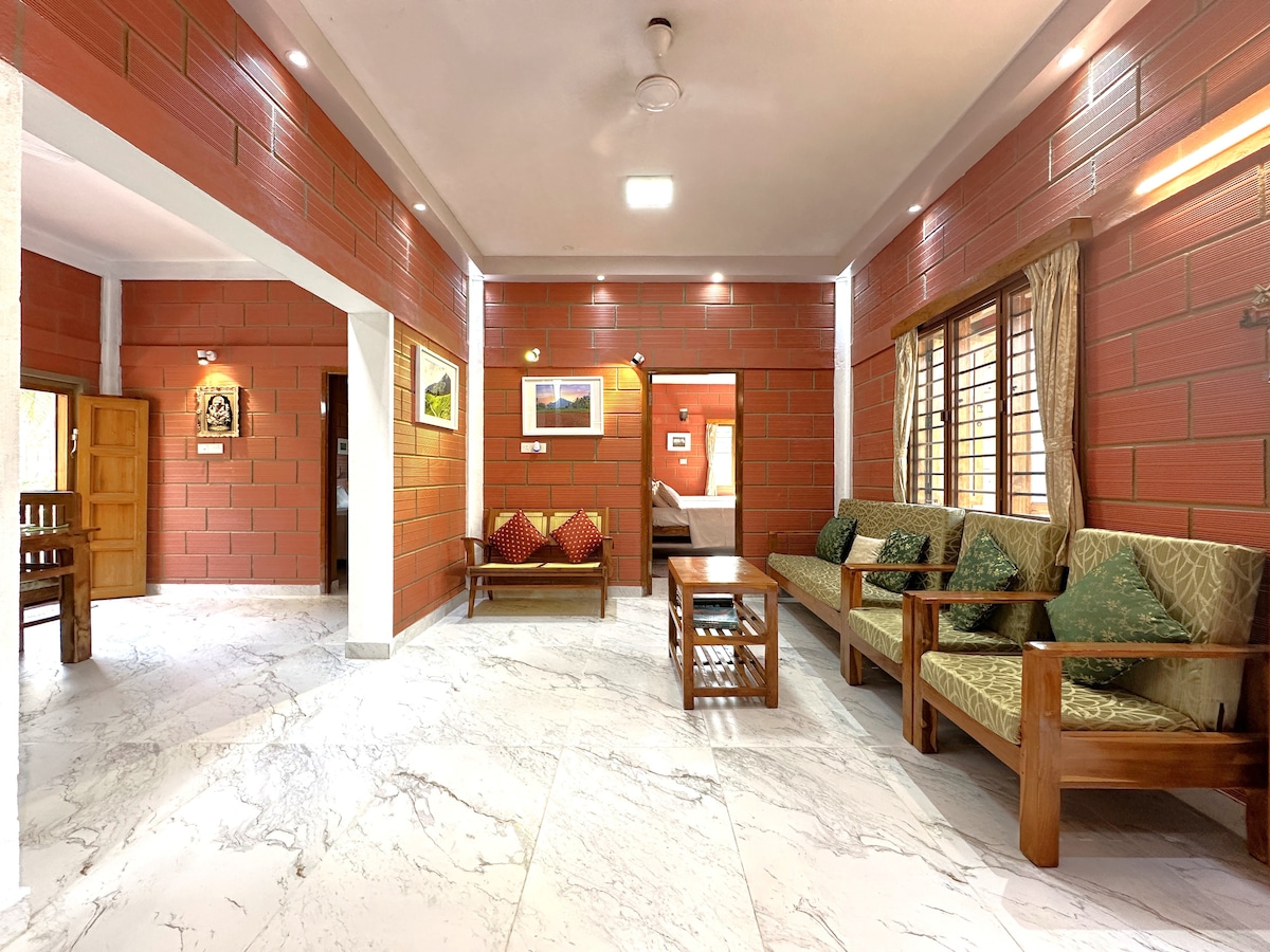 An inviting living area is presented with earthy-toned walls and marble flooring. Comfortable wooden seating is arranged around a central coffee table, while natural light filters in through window treatments. Two entrances lead to additional areas, subtly enhancing the spacious layout.